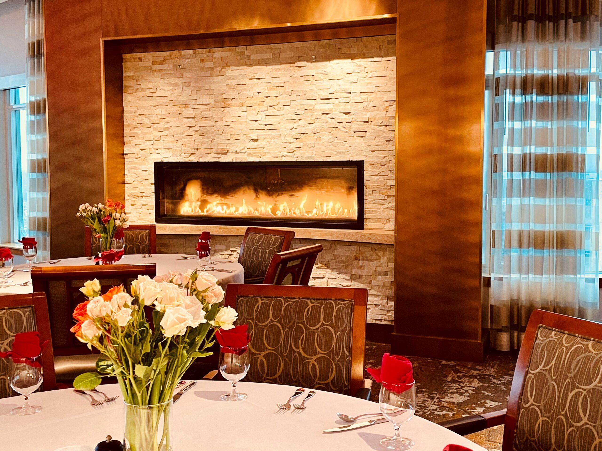 A formal dining area set up for a Valentines Day meal, with vases of flowers at each table and a rose at every seat.